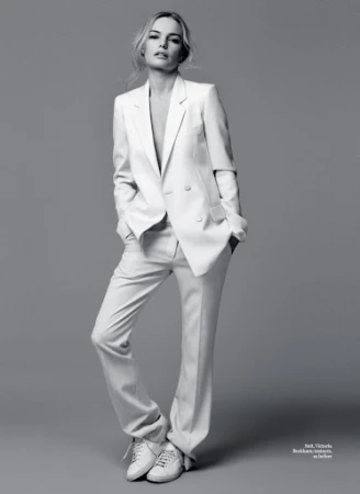 Monochrome portrait of a woman wearing a white suit jacket, matching trousers, and white trainers against a grey background.