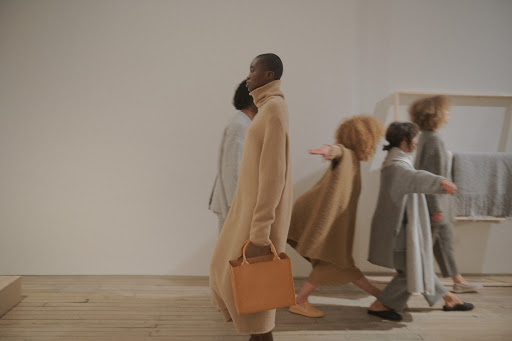 A group of fashionably dressed people walking in studio, an African woman in front carries a tan leather bag; beige tones.