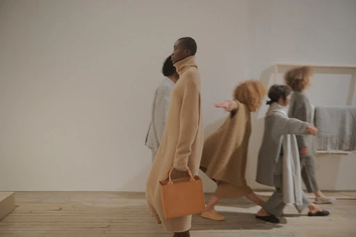 A group of fashionably dressed people walking in studio, an African woman in front carries a tan leather bag; beige tones.