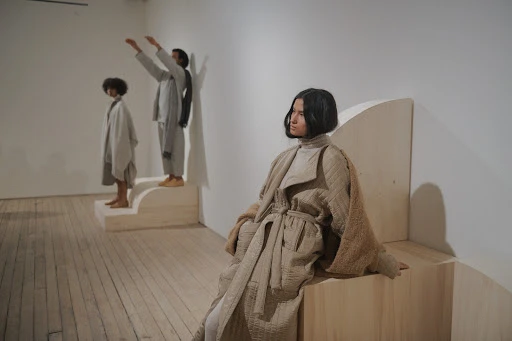 Three people in light colored clothing pose in a minimalist indoor space with wood floors and natural light, showcasing fashion and style.