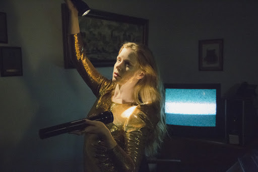 Young woman in a gold sequin dress indoors, shining a flashlight up to the ceiling, with an old television in the background.