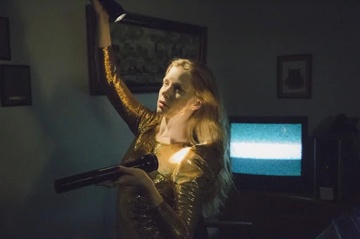 Young woman in a gold sequin dress indoors, shining a flashlight up to the ceiling, with an old television in the background.