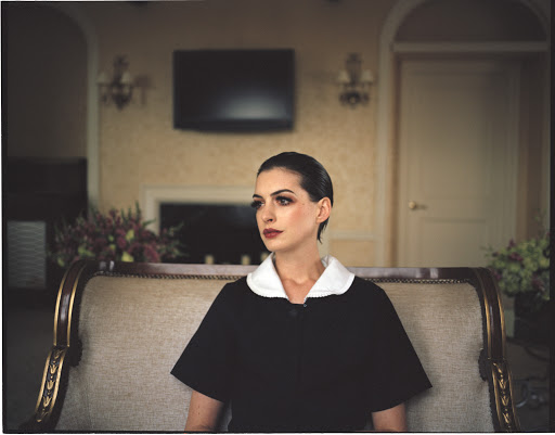 Portrait of a brunette adult woman in a dark dress with white collar, sitting on a gold and tan couch in an indoor setting.