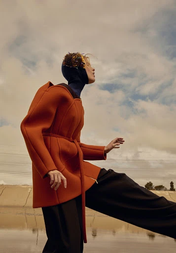 A fashion shot of a blonde model wearing an orange coat and black pants in an outdoor setting with a cloudy sky backdrop.