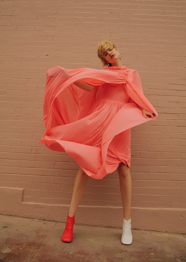 Woman in flowing dress poses against a pink brick wall with red and white ankle boots, backlit with natural light in an outdoor urban setting.