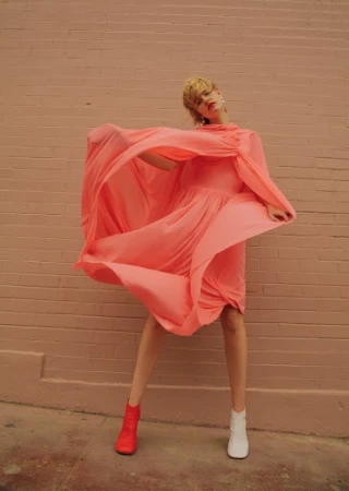 Woman in flowing dress poses against a pink brick wall with red and white ankle boots, backlit with natural light in an outdoor urban setting.