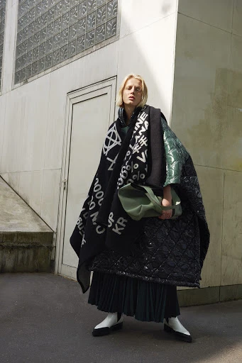 A woman with fair hair stands outdoors wearing a dark, patterned shawl, puffy coat, pleated skirt, boots, and carrying a handbag.