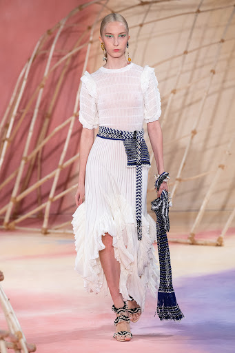Runway fashion show featuring a model in a white textured dress with blue waist accessory and matching heels, posing in a studio setting.
