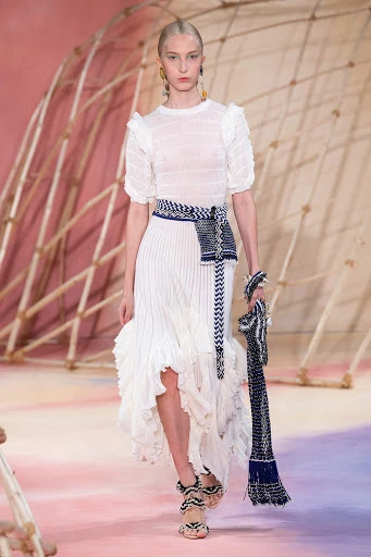 Runway fashion show featuring a model in a white textured dress with blue waist accessory and matching heels, posing in a studio setting.