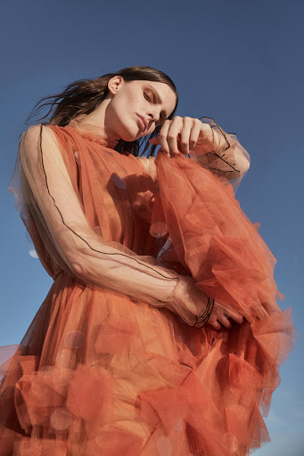 Woman in flowing orange dress posing against a blue sky, fashion editorial-style photography, looking down with a gentle expression.