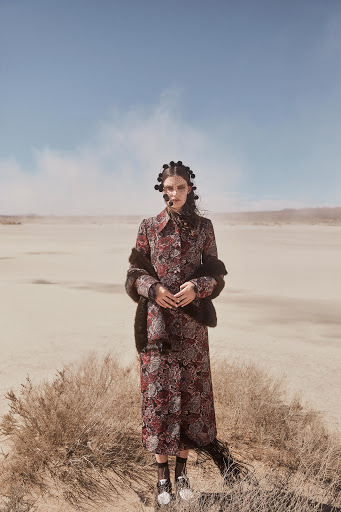 A fashion shot of a woman in a patterned coat and fur stole posing in the desert under a blue sky with some clouds.