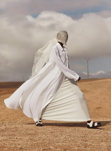 A person wearing a long white dress and head covering walks on a dirt field under a cloudy sky in a fashion editorial style shot.