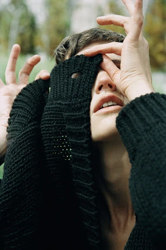 Close-up portrait of an adult woman with her face partially covered by her hands and a black knitted garment outdoors.