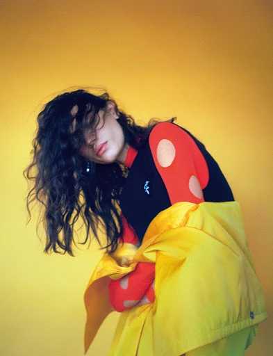 Young woman with curly hair wearing a patterned top and yellow jacket posing against a yellow background in a studio shot.
