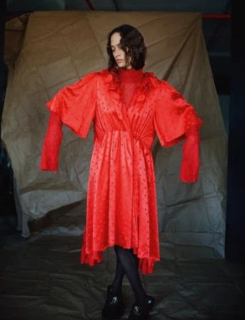 Model in a bright red satin dress with ruffled collar and voluminous sleeves standing against a crumpled paper backdrop.