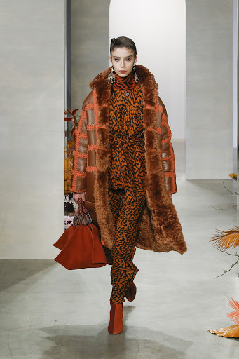Fashion model walks the runway wearing an orange and brown print suit with a large fur coat and carrying an orange handbag.