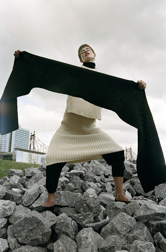Woman in knitwear poses on rocks with long scarf in front of a city skyline under overcast sky. Fashion editorial photography.