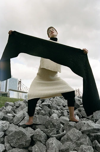 Woman in knitwear poses on rocks with long scarf in front of a city skyline under overcast sky. Fashion editorial photography.