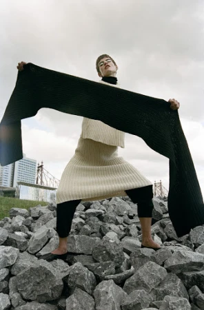 Woman in knitwear poses on rocks with long scarf in front of a city skyline under overcast sky. Fashion editorial photography.