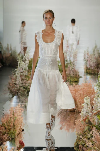 A fair-skinned female fashion model showcases a white dress on a reflective runway during a fashion show with floral accents.
