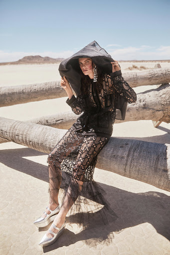 A woman sits on a fallen log in a desert wearing an intricate black hooded lace jacket and skirt, accessorized with statement earrings and silver shoes.