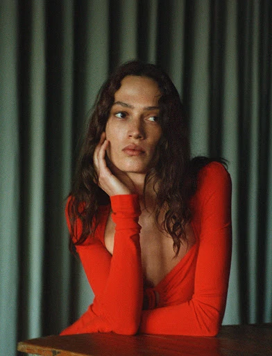Portrait of a woman with long brown hair wearing a long-sleeved red top, resting her face on her hand, sitting at a wooden table.