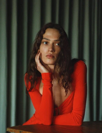 Portrait of a woman with long brown hair wearing a long-sleeved red top, resting her face on her hand, sitting at a wooden table.