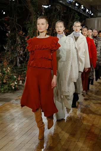Fashion models walk runway wearing elegant earth-toned clothing during an indoor runway show with floral background.