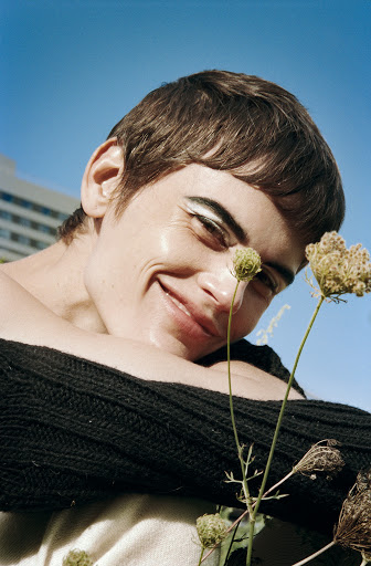 Close-up of smiling adult female looking upwards with white eyeliner, a dark top, and wildflowers in an outdoor setting with a blue sky.