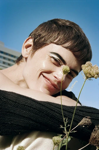 Close-up of smiling adult female looking upwards with white eyeliner, a dark top, and wildflowers in an outdoor setting with a blue sky.