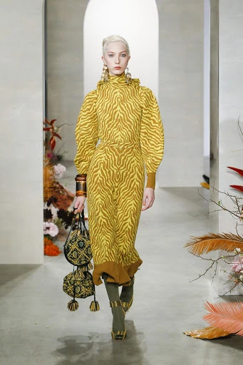 Runway shot of a model wearing a yellow zebra-print dress and carrying a purse, walking in a fashion show with floral decorations.