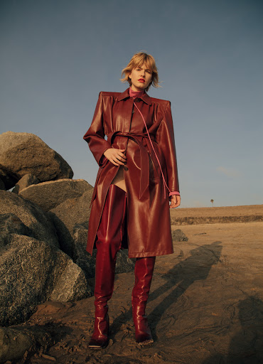Fashionable blonde woman poses in a red vinyl coat and thigh-high boots on a sandy beach with rock formations under a clear sky.