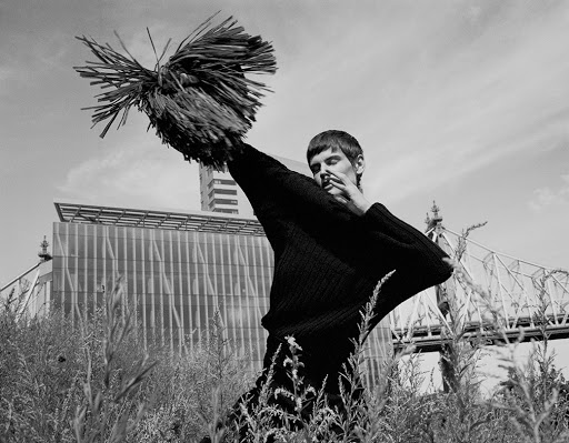 Monochrome fashion shot of a man in a black outfit holding a sculptural straw object, with city buildings and natural grasses in the background.