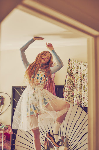 Young woman in floral dress and socks strikes a playful pose, one knee raised, arms above head, indoors with natural lighting, vintage aesthetic.