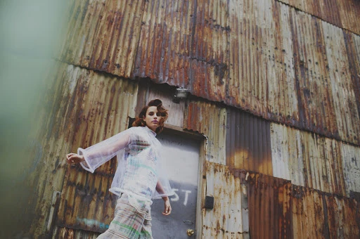 A young woman in a translucent coat and patterned dress stands against a rusty corrugated metal building exterior, styled hair, outdoor setting.