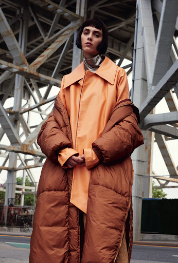 Fashion shot of a brunette adult woman in a long orange coat and brown puffer jacket standing under a metal bridge outside in daytime.