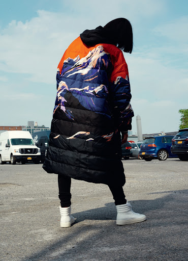 Woman poses in a mountain-print puffer coat, black leggings, and high-top sneakers against an urban parking lot backdrop under a cloudy sky.