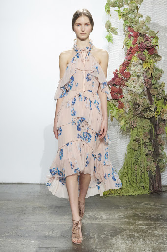 Woman walks runway in light pink floral dress with strappy heels against floral and white backdrop in indoor fashion show setting.
