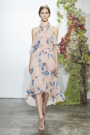 Woman walks runway in light pink floral dress with strappy heels against floral and white backdrop in indoor fashion show setting.