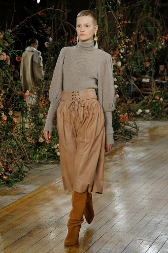 Runway fashion show featuring a woman in a grey sweater, tan skirt, and boots, walking on a wooden floor with a floral backdrop.