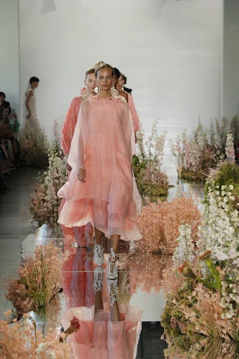 Fashion show runway featuring models in light pink sheer gowns walking amidst floral arrangements in a brightly lit indoor space.