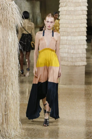 Runway fashion show featuring a model in a color-blocked halter dress, strappy sandals, and tassel earrings, surrounded by other models.