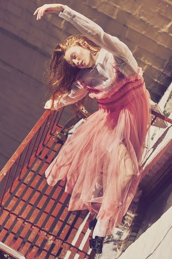 Young woman in a pink tulle dress poses on a metal fire escape with white socks and black shoes, captured in an urban setting with natural light.