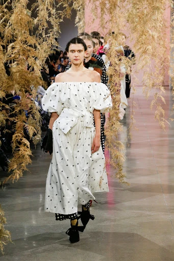 Fashion show runway featuring a female model in a white, off-the-shoulder dress with black accents, walking towards the viewer indoors.