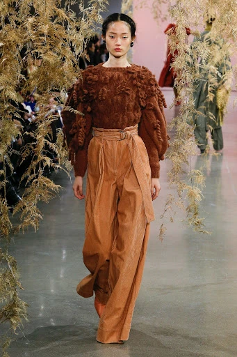 Runway fashion: A model walks the catwalk wearing a brown textured sweater and tan wide-leg pants, framed by floral accents.