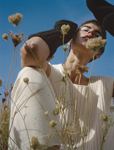 Person wearing a white top posing in a field of plants against a blue sky, with a black glove or mitten partially visible.