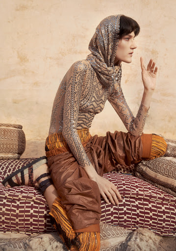 A woman in patterned top and headscarf sits among patterned cushions with a neutral backdrop in a studio setting, creating a unique fashion image.