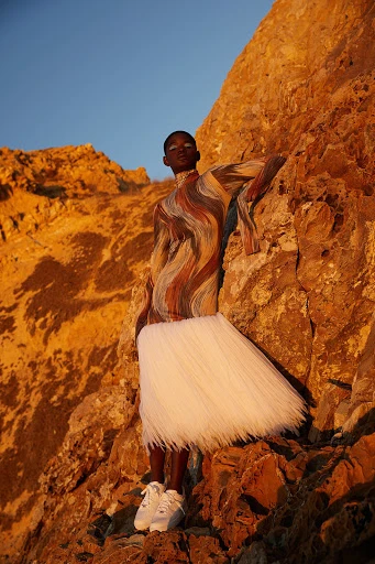 A woman in a patterned top and white skirt poses on a sunlit rock formation against a clear sky in an outdoor fashion shot.