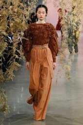 Model walking a runway wearing a textured brown sweater and pleated high-waisted trousers framed by hanging dried foliage