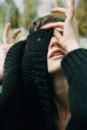 Close-up portrait of a person partially concealing their face with a textured black knitted sweater and raised hands.